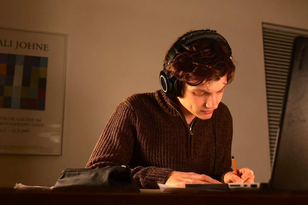 Julius Fischer beim Arrangieren mit Stift und Papier vor einem Laptop. Er trägt einen braunen Pullover und Kopfhörer.